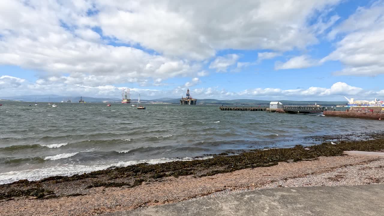 Wide shot of choppy sea with sailboats and fishing boats near Cromarty, Scotland. Cloudy sky, bright daylight, gentle camera pan, natural coastal environment
