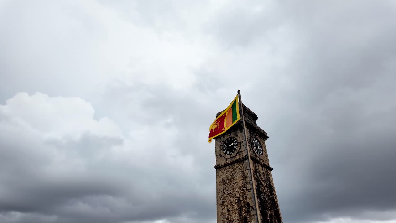 The historic Galle Fort Clock Tower prominently displays the Sri Lankan flag, fluttering in the wind under a moody, cloudy sky.