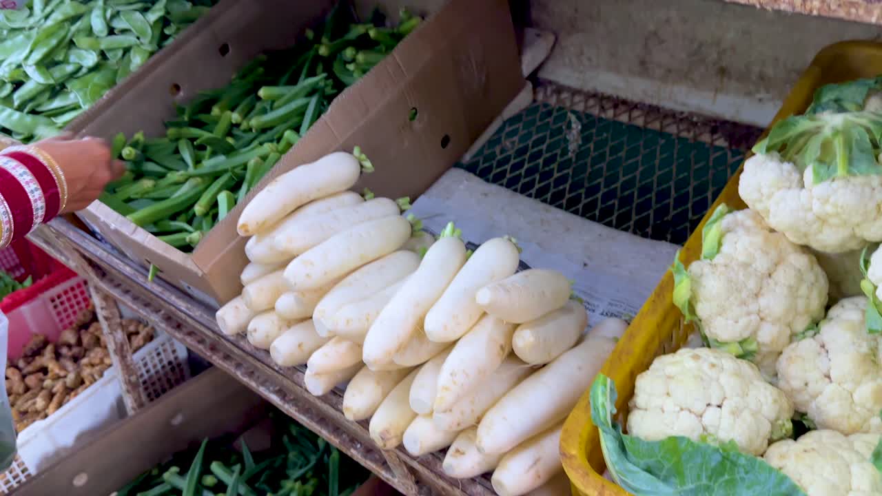 Person chooses okra beside cauliflower and daikon under natural daylight at outdoor vegetable market