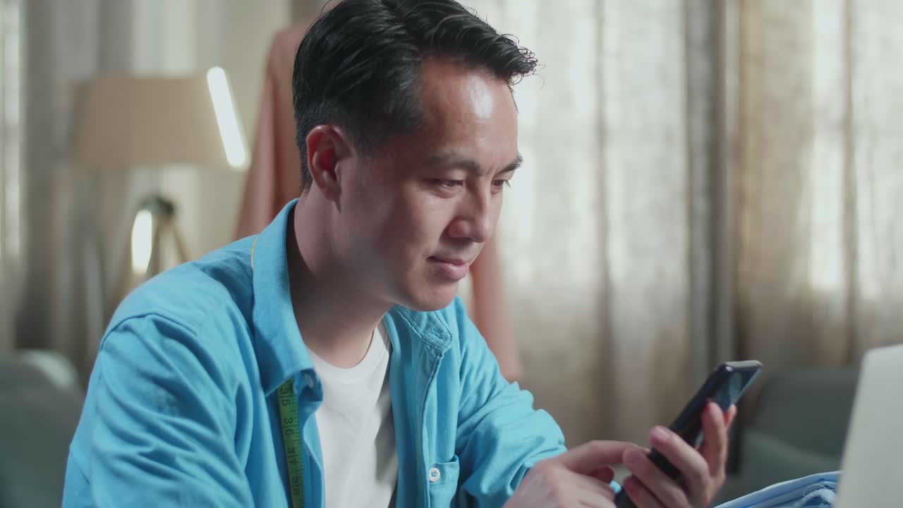 스튜디오에서 노트북 컴퓨터에서 옷을 디자인하는 동안 스마트 폰을 보는 바느질 기계와 함께 아시아 남성 디자이너의 클로즈업