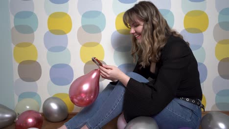 mujer joven sentada alrededor de globos y sosteniendo un teléfono en la mano contra un fondo colorido