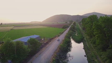 aerial shot as car drives down rural road by creek in between farmland, 4k