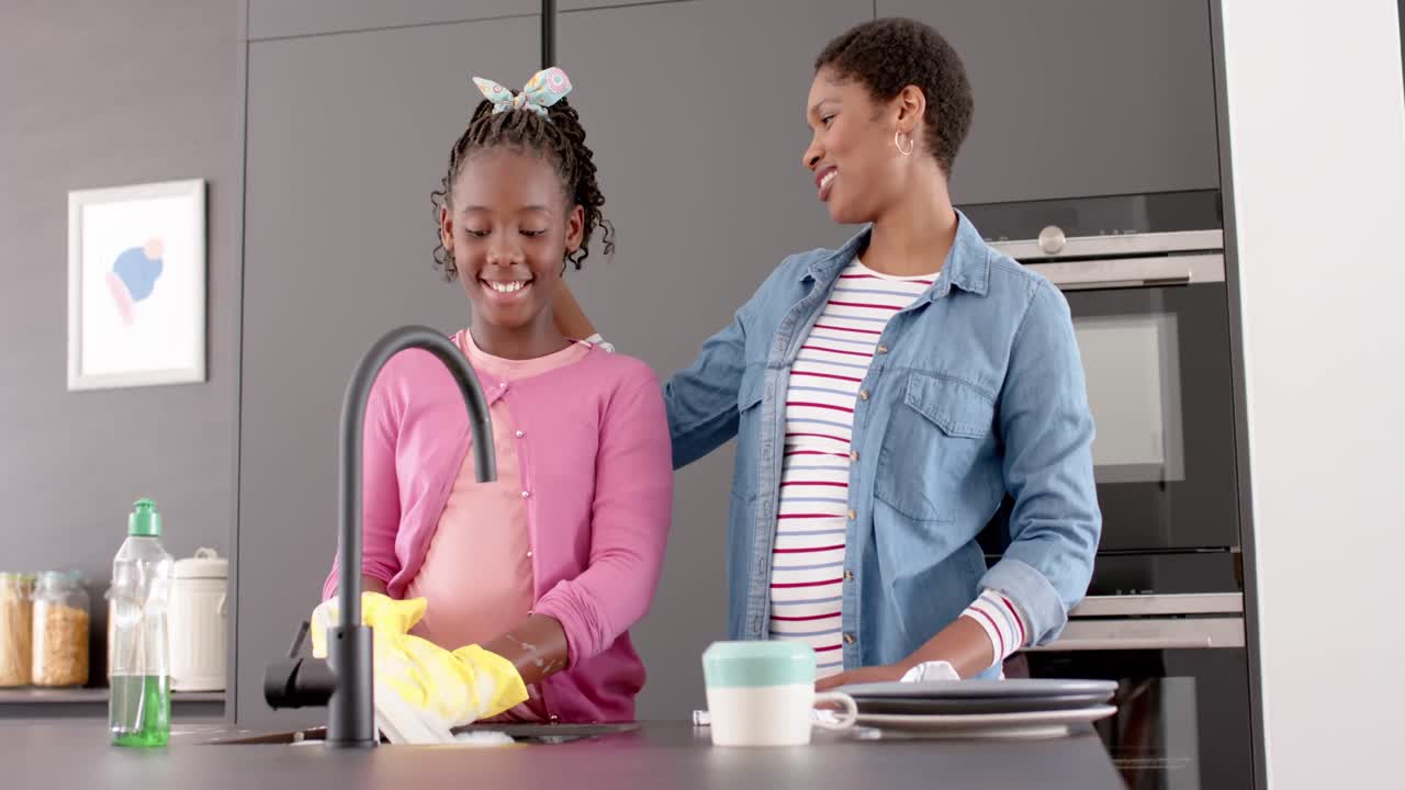 Happy african american mother and daughter washing dishes and embracing in kitchen, slow motion