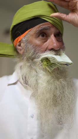 video vertical de primer plano de un anciano sikh con barba atando tela para el turbante y usando aguja salai contra un fondo plano