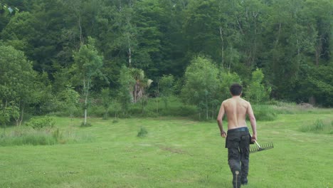 wide shot topless man walking up field with rake