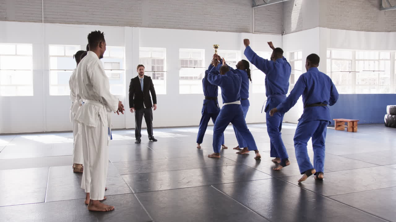 Judo team celebrating victory with trophy in dojo, coach observing proudly