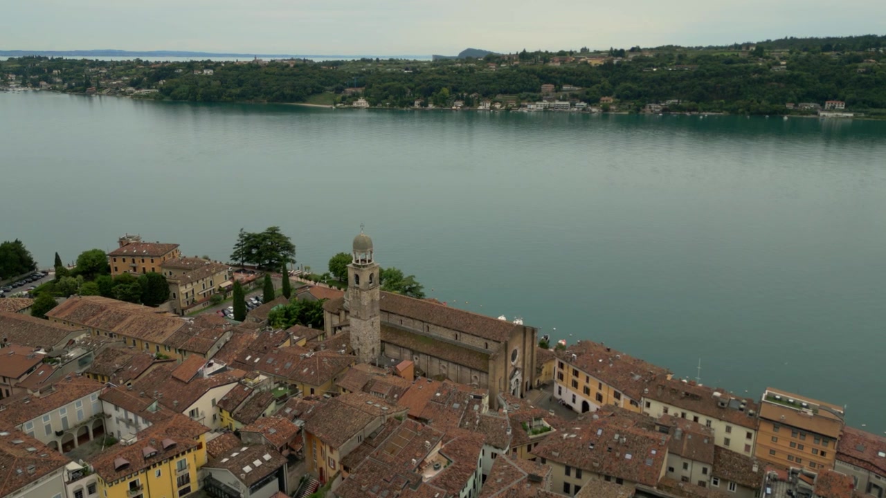 Vista aerea che scivola sopra i tetti storici di Salò con lago e colline oltre.