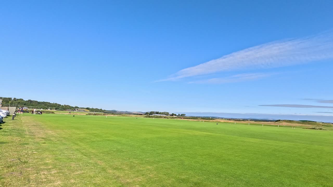 Camera pans right, revealing parked cars beside green golf course under clear blue sky