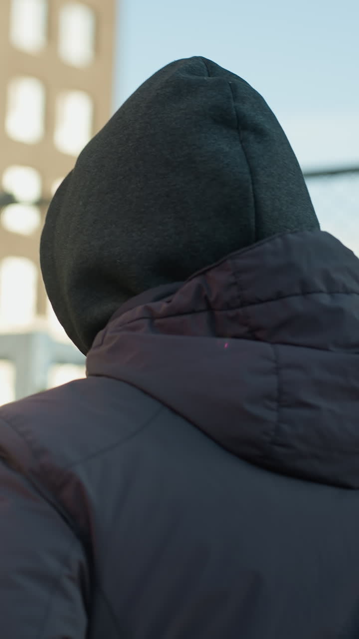 atleta con capucha y gorra corriendo en una arena deportiva con poste de portería en el fondo y vista borrosa de personas caminando, rodeado de arquitectura urbana