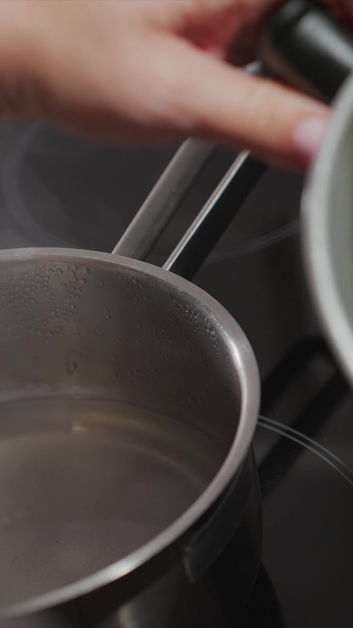 Close up of hand carefully pouring liquid from white container into stainless steel pot on induction cooktop. water level rises steadily creating condensation droplets inside metal vessel