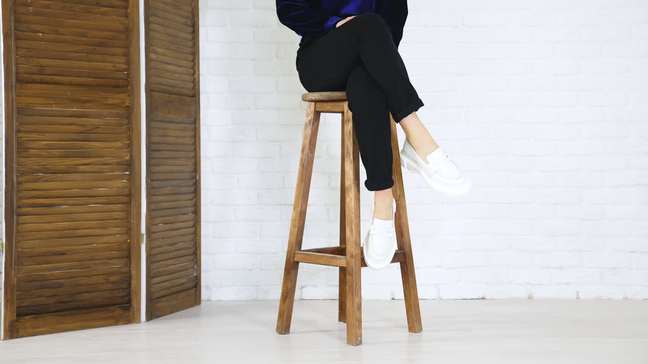 Unrecognized woman sitting on the stool showing her new shoes. Presenting white modern design footwear at white wall backdrop.