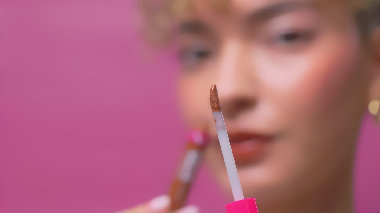 Close up of woman applying brown lip gloss in slow motion with soft pink background
