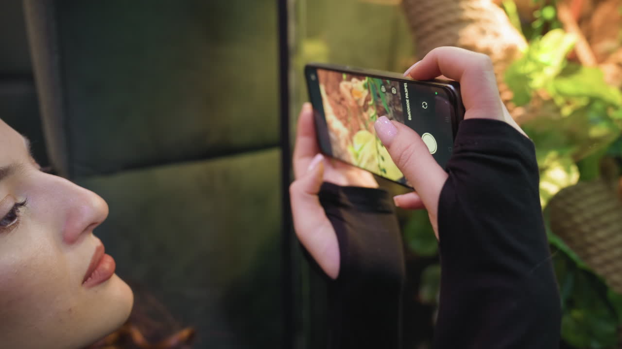 Closeup view of woman holding black phone with pink acrylic nails while capturing photo near vibrant green terrarium. Bright natural lighting enhances leafy texture and colors in background scene