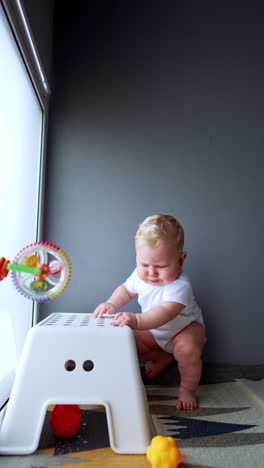 beautiful blond baby standing at the chair smiling adorably. kid sits down on the floor pulling a chair closer. vertical video.