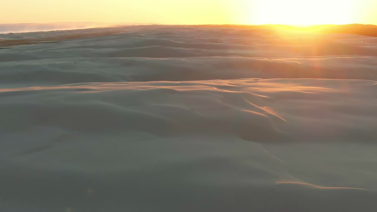Flyover of dunes in Australia next to the ocean as the camera tilts and pans to reveal a golden sunset on the horizon