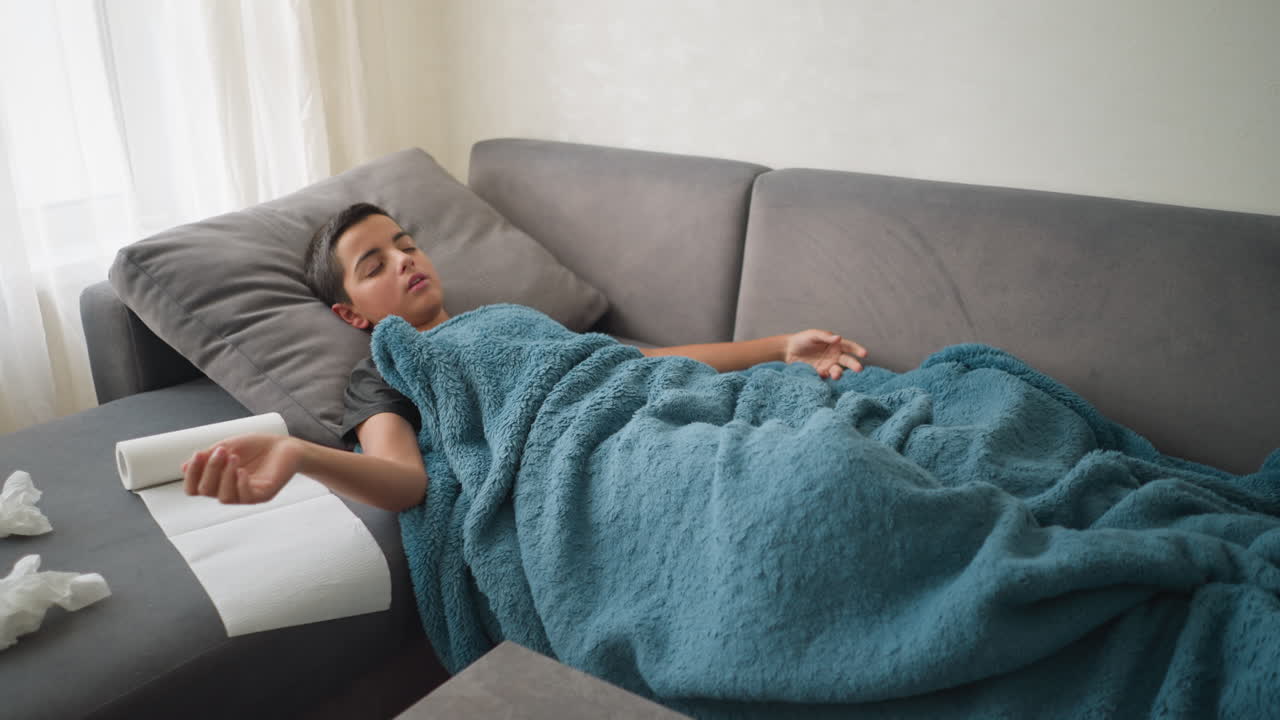 Aerial view of scattered room with boy lying on couch under blanket, adjusting while glass cup and tissues are on the table, soft, cozy atmosphere with a sense of illness or rest