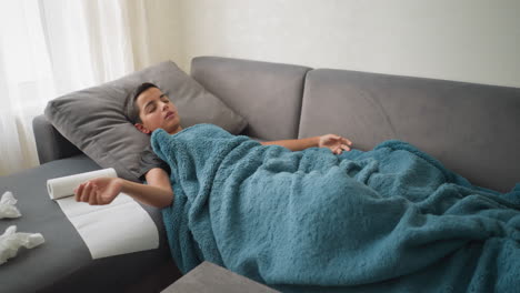 aerial view of scattered room with boy lying on couch under blanket, adjusting while glass cup and tissues are on the table, soft, cozy atmosphere with a sense of illness or rest