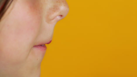 a close-up captures a child taking a big bite from a berliner-style donut with yellow frosting and chocolate flakes, held in a hand with orange nail polish against a vibrant yellow background.
