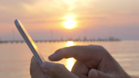 man typing on smartphone by sea at sunset