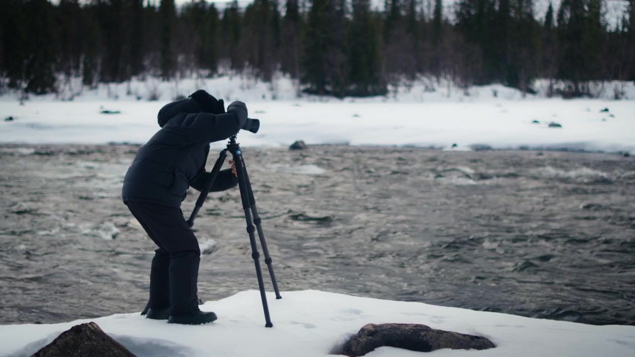 En indpakket kameramand læner sig over et stativ på en sneklædt flodbred og filmer hurtigt, koldt vand med mørk skov i baggrunden.