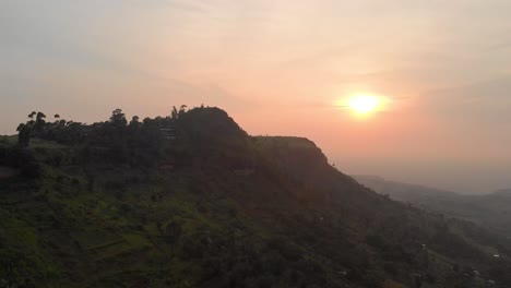 Aerial-shot-orbiting-around-a-large-hill-in-the-mountains-of-tropical-East-Africa-while-the-orange-sun-sets-over-the-land