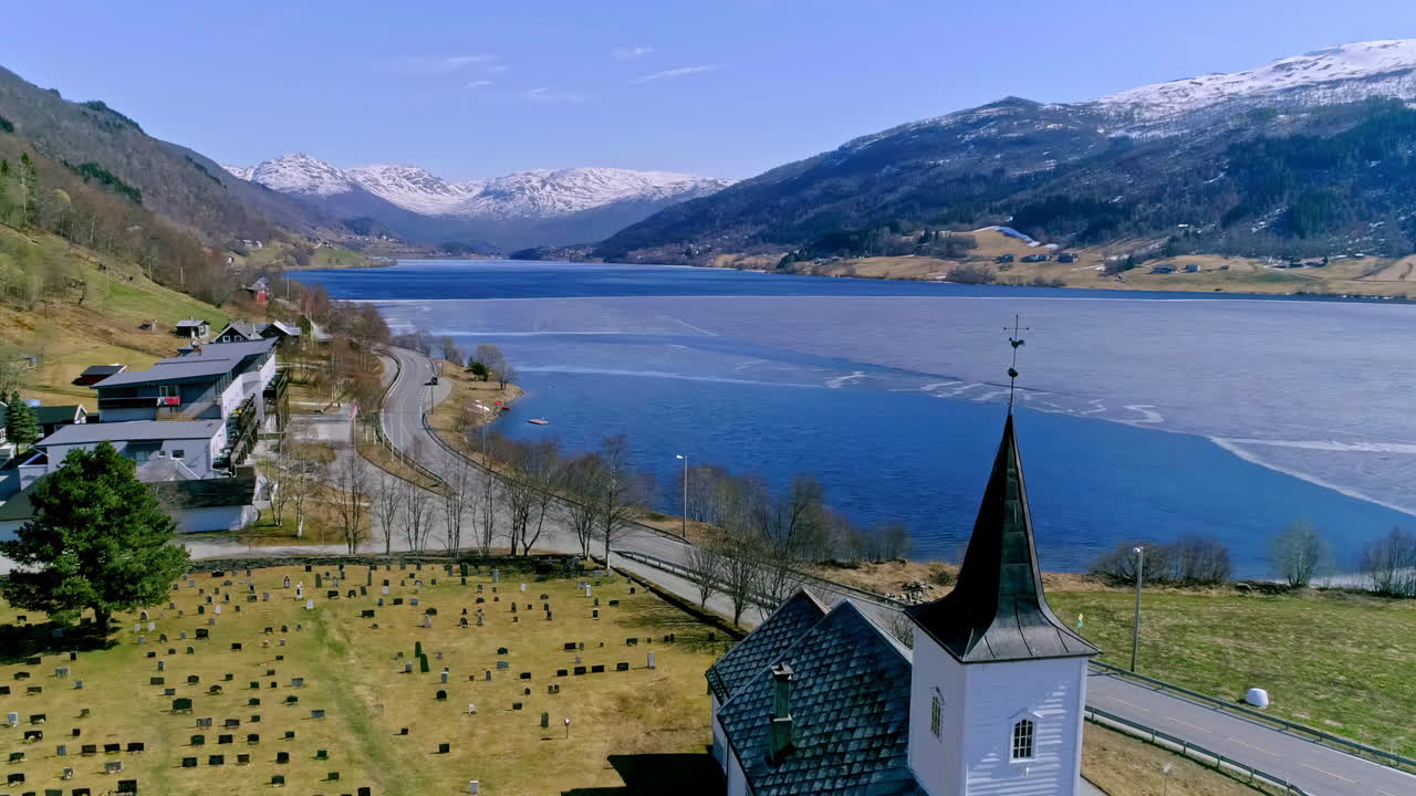 toma aérea de un lago nevado junto a un valle verde con una iglesia con una veleta y un cementerio
