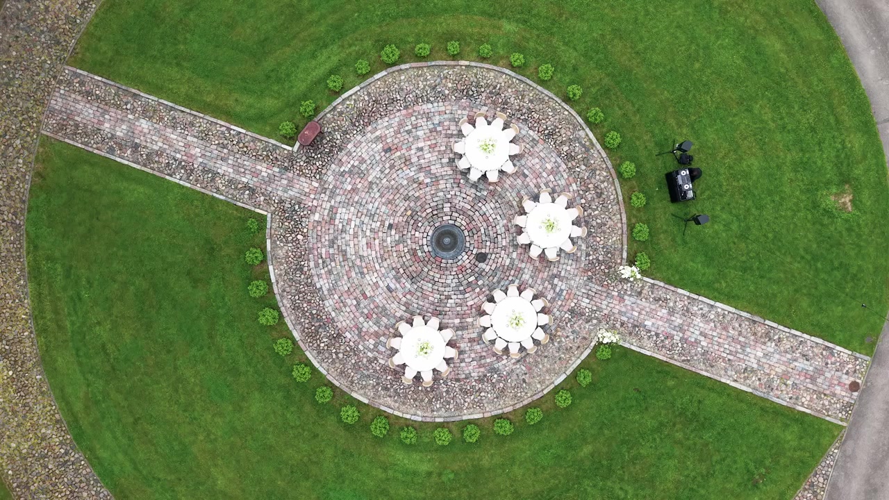 Imagens aéreas de um caminho circular simétrico de tijolos com quatro grandes esculturas florais brancas, rodeado por um exuberante relvado verde à luz do dia. A tomada aérea oferece uma composição clara e geométrica.