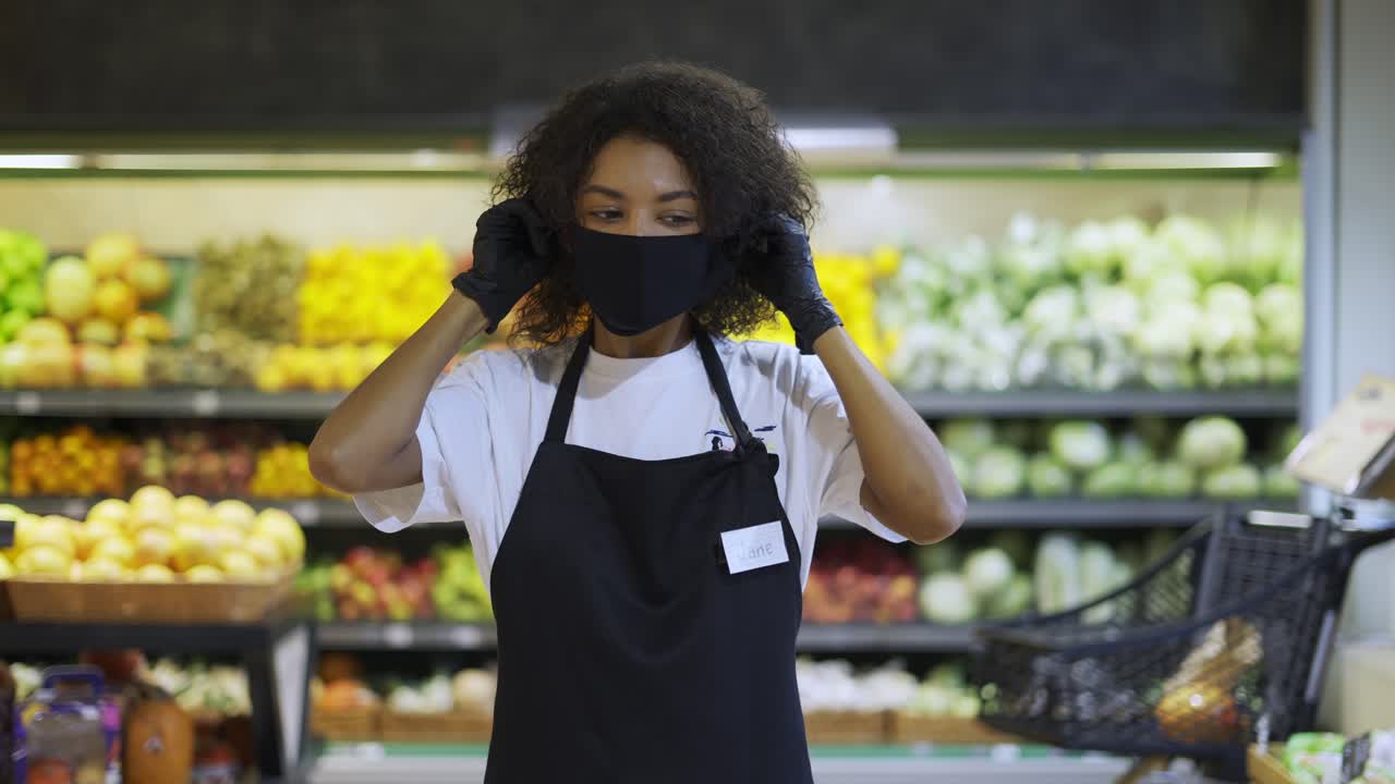 Retrato de funcionária afro-americana feliz em pé no supermercado, colocando máscara de proteção.