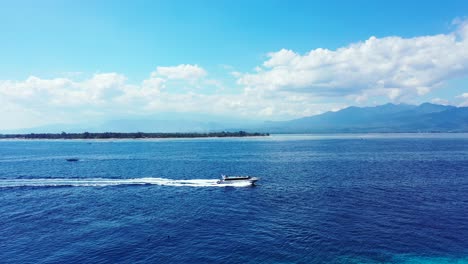 Speed-boat-sailing-across-vibrant-blue-sea-surface-leaving-shore-of-tropical-island-,-deep-ocean-on-a-bright-morning-sky-with-clouds