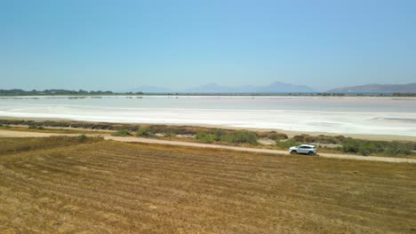 Vista-Aérea-De-Un-Automóvil-Blanco-Conduciendo-Por-El-Camino-De-Tierra-Cerca-Del-Salar-De-Igroviotopos-Alikis-En-Kos,-Grecia