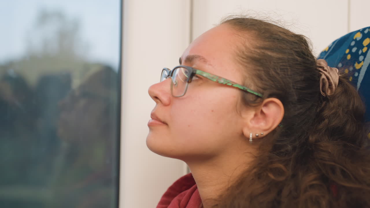 Young woman on train at dusk listening earbuds, art student reviewing ideas, glances toward blurred skyline, warm interior light, subtle glass reflection, contemplative expression, soft playlist