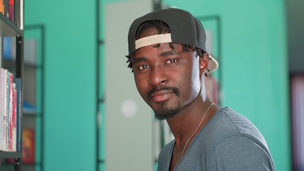 Side view of student in casual cap and gray shirt standing near book shelf in bright library, turning head to look at camera with focused expression against blurred background