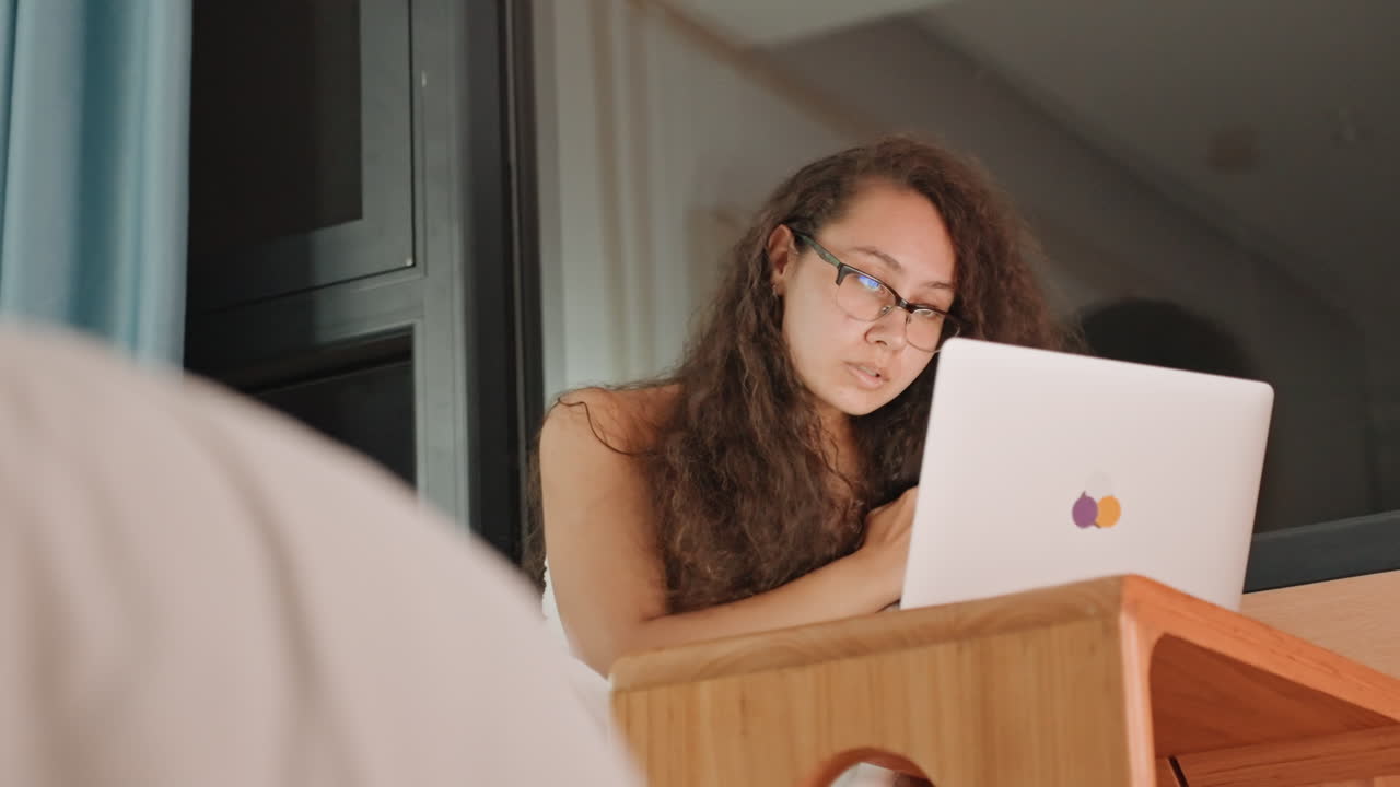 Mulher estudando em laptop em quarto de hotel, cena de escrivaninha focada com pequena mesa de madeira, óculos, janela com cortina e paisagem urbana noturna, atmosfera concentrada transmitindo estudo, pesquisa e criatividade