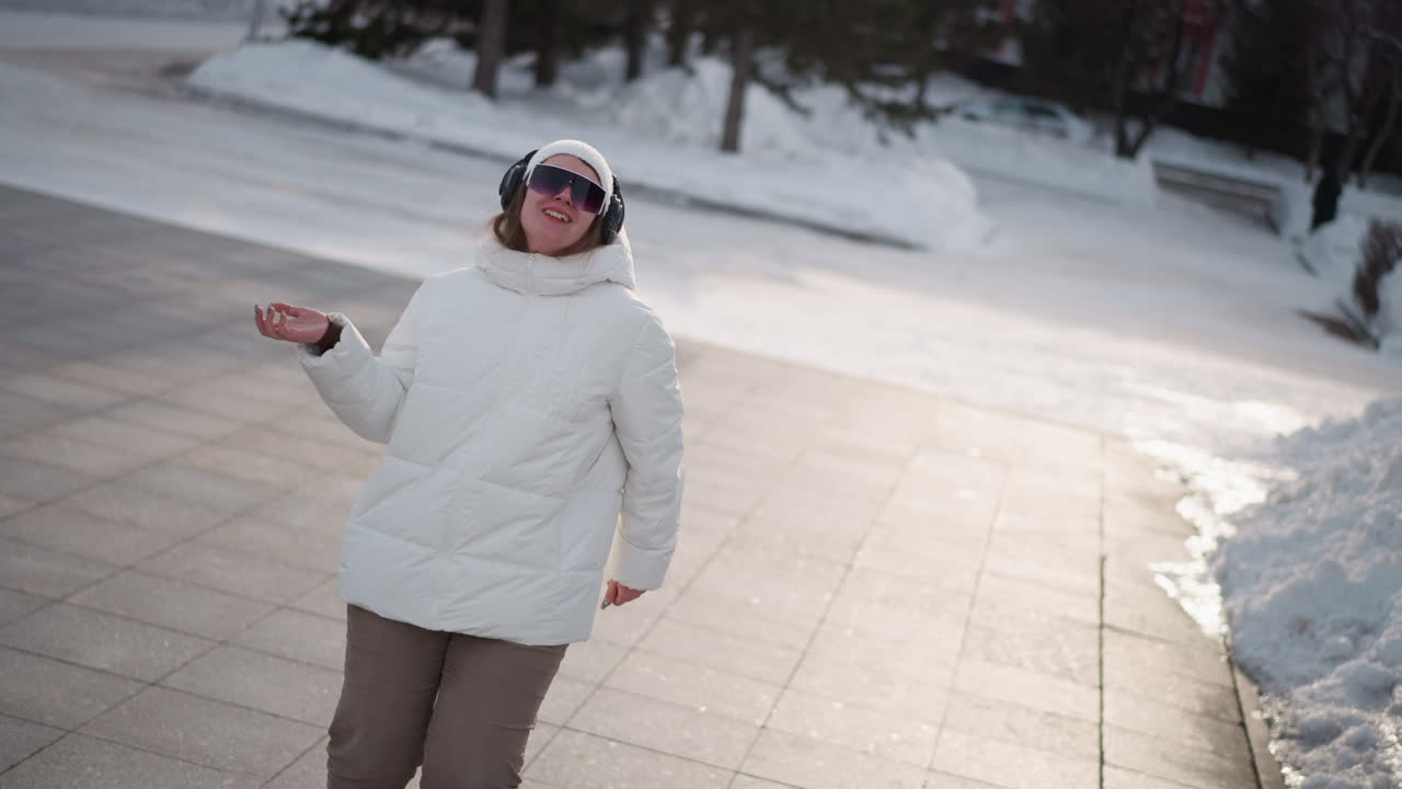 Free spirit woman joyfully spinning with excitement while dancing in white puffer jacket, beanie, goggles and headphones on snow dusted urban plaza, moving arms and body rhythmically