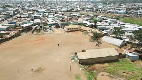 poor school children moving out of school, poor african ethnical people passing over the brown field, poor sheet settlement surrounding the football pitch in kibera nairobi kenya