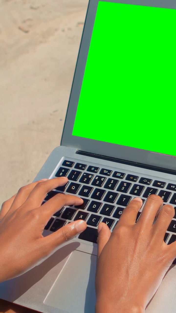 Video verticale: Mani adulte iniziano a digitare, premendo i tasti del laptop su una spiaggia sabbiosa vicino a uno schermo verde.
