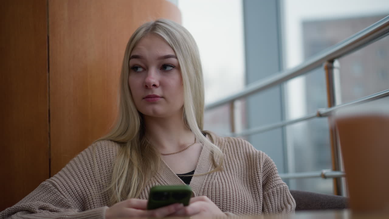 молодая женщина с цепью на шее сидит и держит телефон, выглядит задумчиво с размытым видом на город, видимым через окно, современная, расслабленная атмосфера в городской обстановке