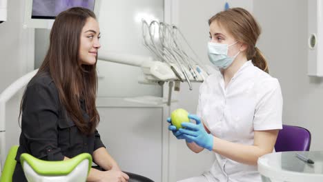 Young-female-dentist-offering-green-apple-to-happy-and-healthy-patient-after-the-procedures