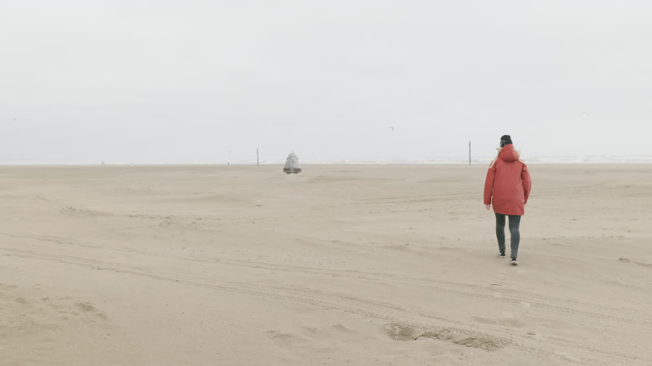 vista trasera de una chica con una chaqueta roja caminando por la orilla arenosa de la playa de ijmuiden en una mañana fría en los países bajos