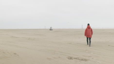 back view of a girl wearing a red jacket walking on the sandy shore of ijmuiden beach on a cold morning in the netherlands