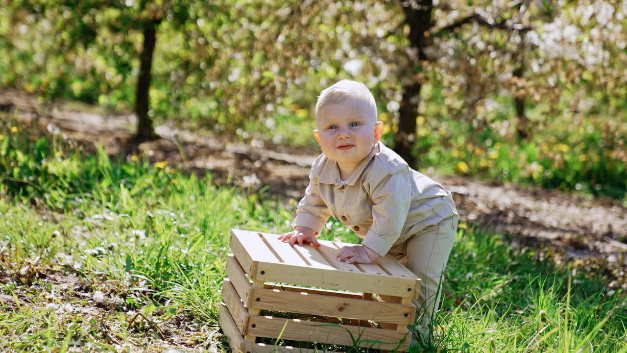 Adorable blond baby boy wearing beige romper stands leaning on the wooden crate. Lovely smiling child looks around the beautiful spring garden.