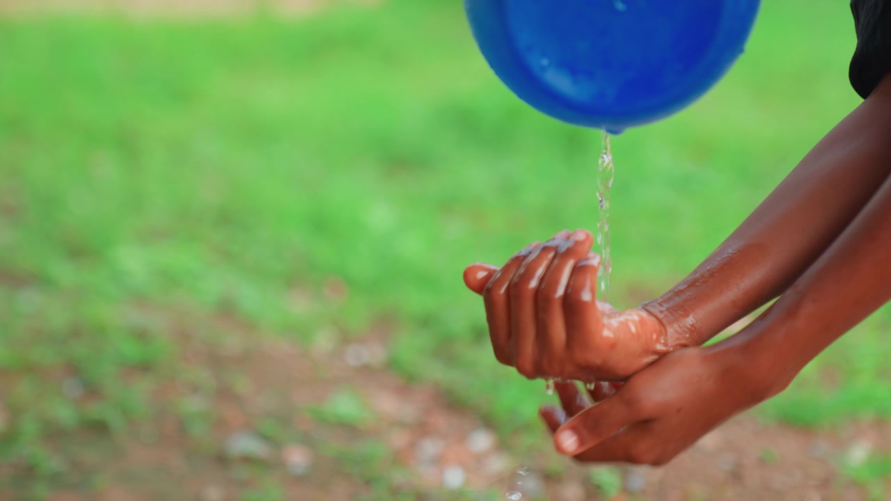 Kind giet water uit blauwe teil, handen komvormig onder de straal, speelse reinigingsbeweging, landelijke tuinomgeving, druppels verspreiden over handpalmen, eenvoudig hygiëneritueel, natuurlijke groene ondergrond, verfrissend