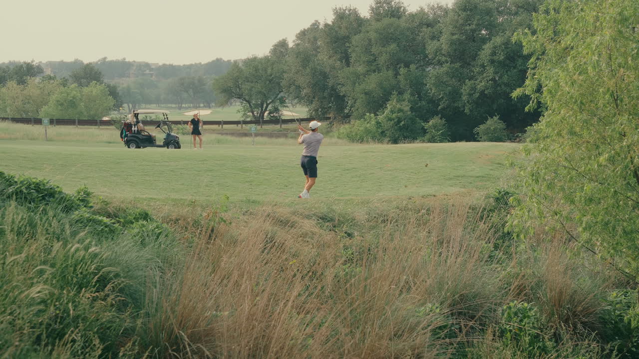 A male golfer hits a fairway shot out of the rough with a divot flying. His female partner stands beside a golf cart in the background, watching. A dynamic two-player golf moment in real time.