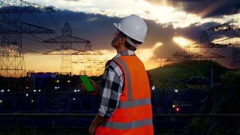 back view of asian male engineer with safety helmet working on a green screen smartphone and looking around while standing near high voltage tower