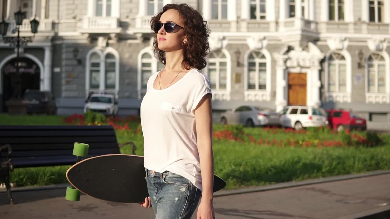 hermosa mujer joven con gafas de sol caminando por la calle de la ciudad vieja sosteniendo su longboard por la mañana durante el amanecer