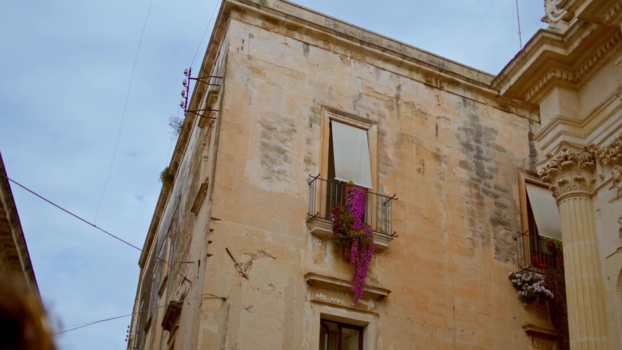 Buganvillas moradas caen en cascada desde un balcón de un edificio histórico de piedra caliza en Ostuni, Brindisi, Puglia, Italia, destacando la arquitectura mediterránea, el encanto urbano y el carácter residencial local.