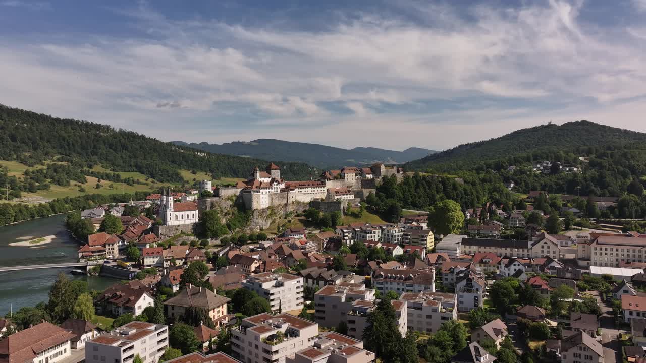 An aerial view showcases the picturesque town of Aarburg, Switzerland, with its historic castle perched atop a hill, overlooking the Aare River. The town's buildings are nestled in a valley