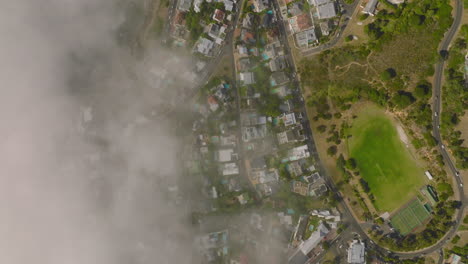 Fly-above-coastal-city,-limited-visibility-due-fog-rising-from-sea.-Houses,-streets-and-sports-areas-in-urban-neighbourhood.-Cape-Town,-South-Africa