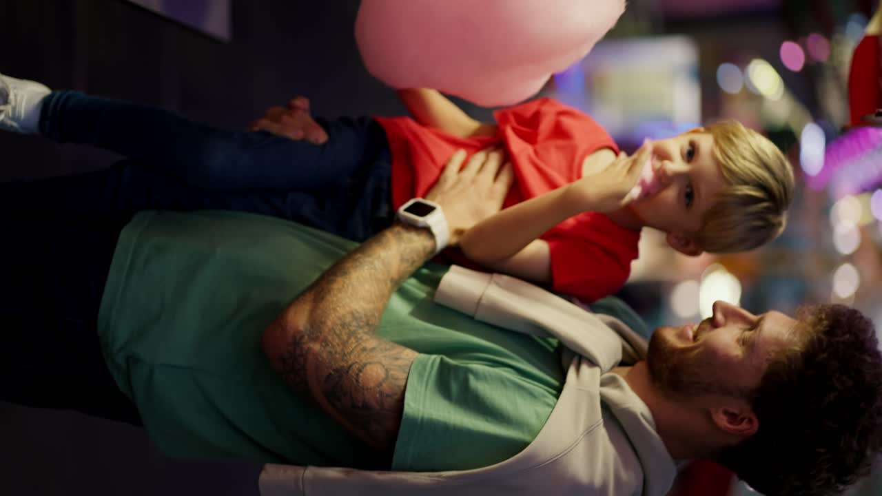 Vertical video: Little blond boy in a red T-shirt eats pink cotton candy with his dad in a Green T-shirt. Father and son going to amusement park