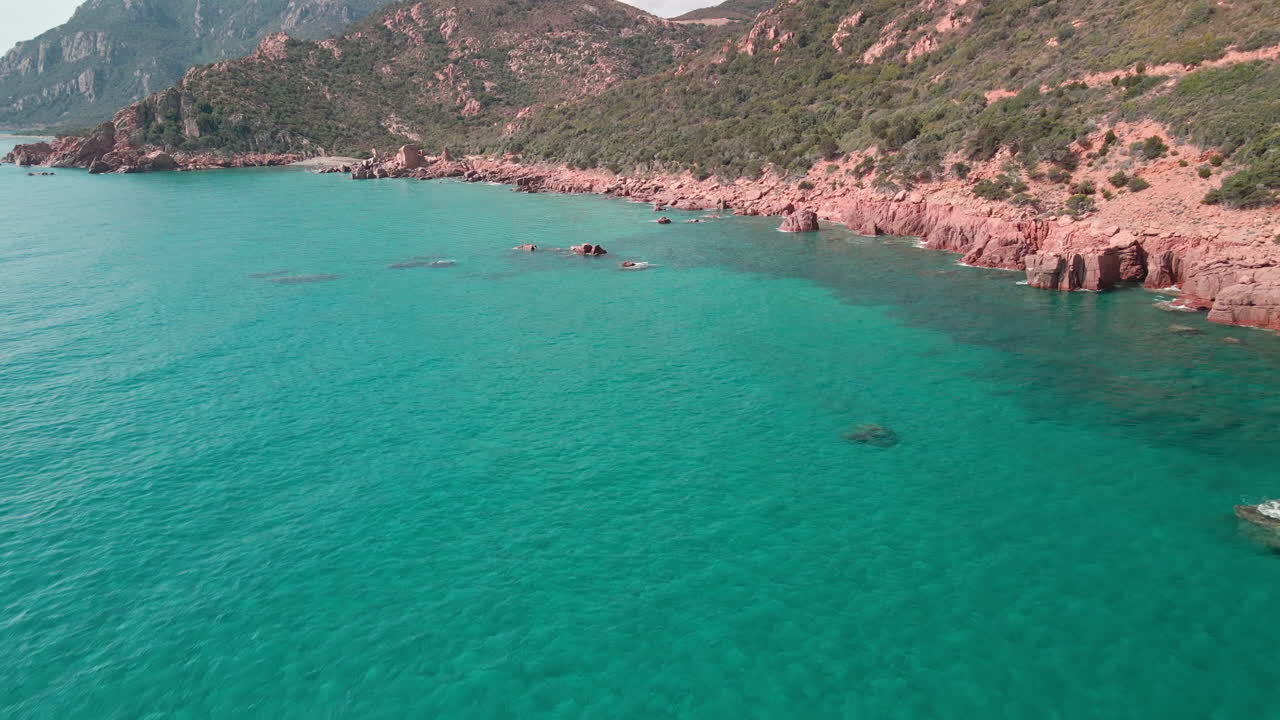 이탈리아 사르데냐의 바위에 부서지는 파도와 함께 열대 푸른 바다의 붉은 산과 푸른 초목의 해안 근처에서 무인 항공기 보기 - 공중 촬영